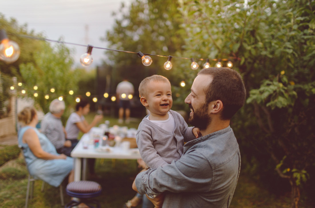 Eine Gruppe von Leuten feiert in einem mit Lichterketten erleuchteten Garten. Ein Mann trägt ein Kleinkind. 